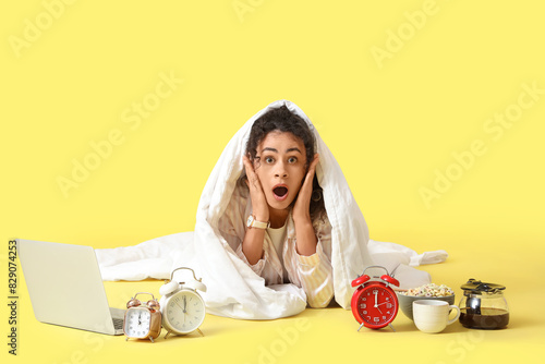 Overslept young African-American woman with blanket, breakfast, laptop and alarm clocks on yellow background