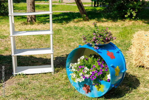 A blue barrel with flowers in it sits on the grass next to a white shelf, decorative drum