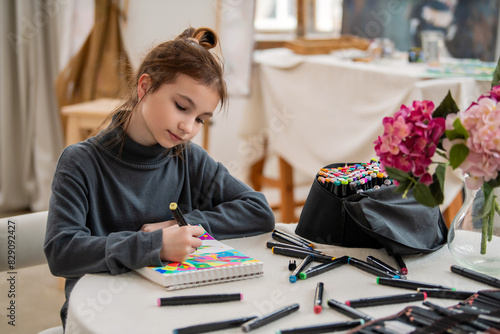 A girl is sitting at a table with a notebook and a bag of markers.