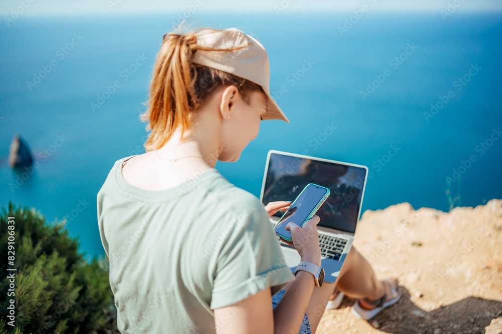 A woman is sitting on a rock by the ocean, using her laptop and cell phone
