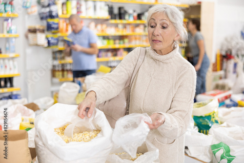 Interested elderly woman buying bulk products in grocery store  standing near bags of various grains  using small scoop to fill polybag with dried corn