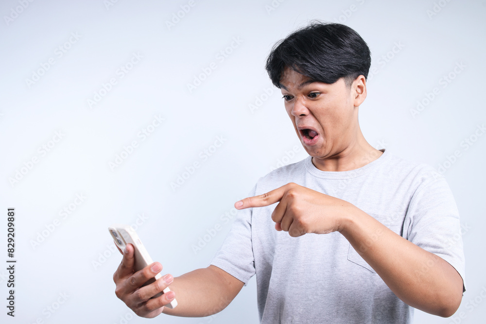 Potrait Of Angry Young Asian Man Scolding And Pointing To Smartphone Isolated On White Background Potrait Of Angry Young Asian Man Scolding And Pointing To Smartphone Isolated On White Background