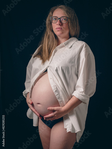 A pregnant woman in a white shirt reveals her bare belly against a dark background. She gently holds her belly, signifying loving connection to her unborn child. Beauty and anticipation of motherhood.