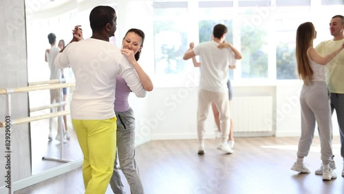 Positive adult black man in bright yellow tracksuit confidently waltzing with fit brunette during group slow ballroom dancing class in choreography studio. High quality 4k footage photo