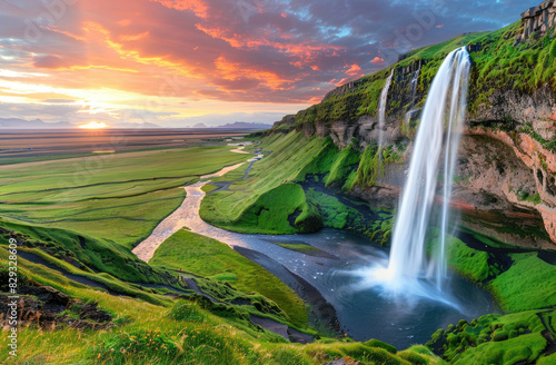 The majestic vaulting waterfall of the circular shape is located in an enchanting valley with lush green grass and vibrant colors of sunset sky.