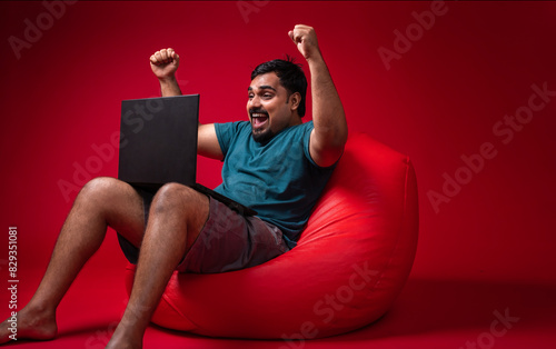 Excited young Indian man sitting on bean bag and looking at laptop, Successful happy person using laptop, Big offer concept background
