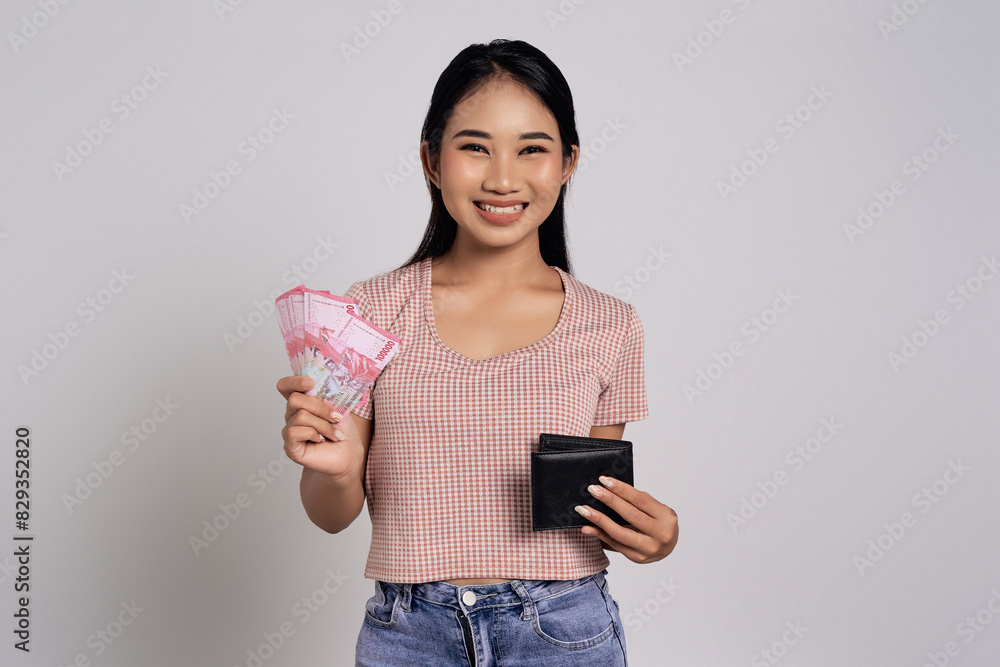 Smiling happy beautiful young Asian woman in a casual t-shirt holding wallet and showing cash money in rupiah banknotes isolated on white background. Financial business concept Smiling happy beautiful young Asian woman in a casual t-shirt holding wallet and showing cash money in rupiah banknotes isolated on white background. Financial business concept
