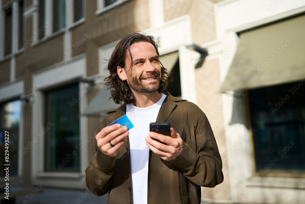 Smiling handsome man using smart phone doing online shopping through credit card on sunny day
