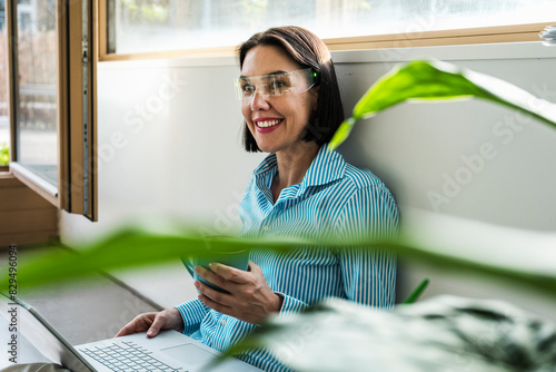 Smiling businesswoman wearing smart glasses sitting with laptop and coffee cup at office photo