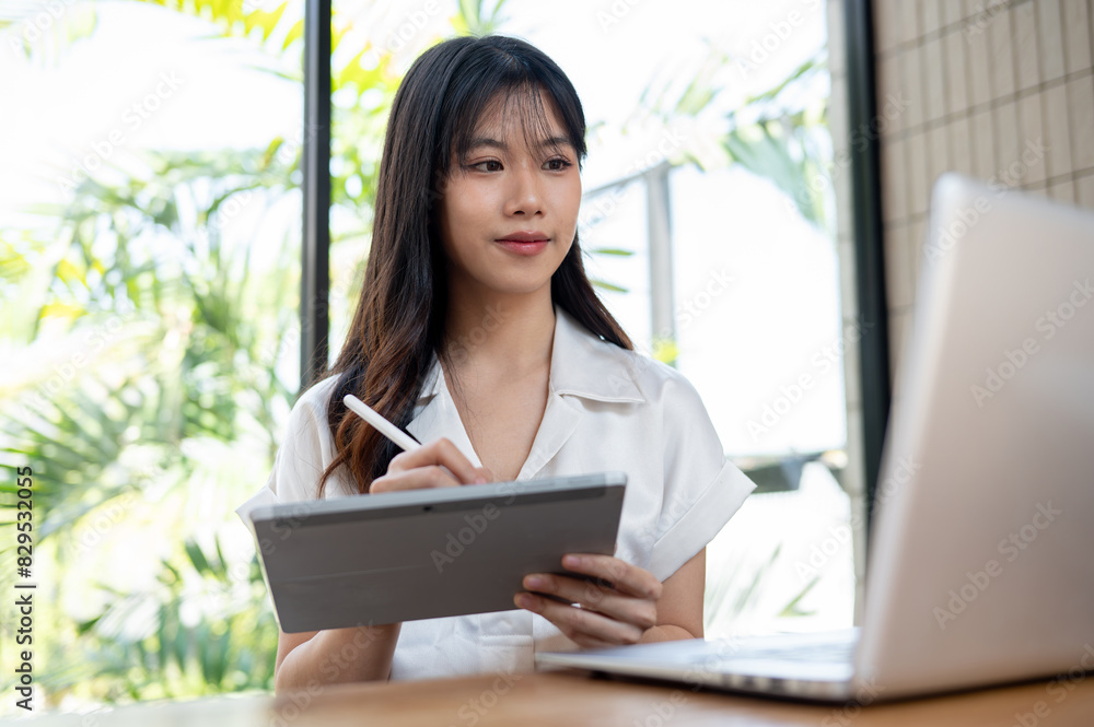 An attractive Asian businesswoman multitasks, using her digital tablet and laptop computer in office An attractive Asian businesswoman multitasks, using her digital tablet and laptop computer in office