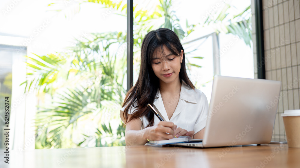 A beautiful Asian woman is listing her ideas in a notebook while browsing the internet on her laptop A beautiful Asian woman is listing her ideas in a notebook while browsing the internet on her laptop