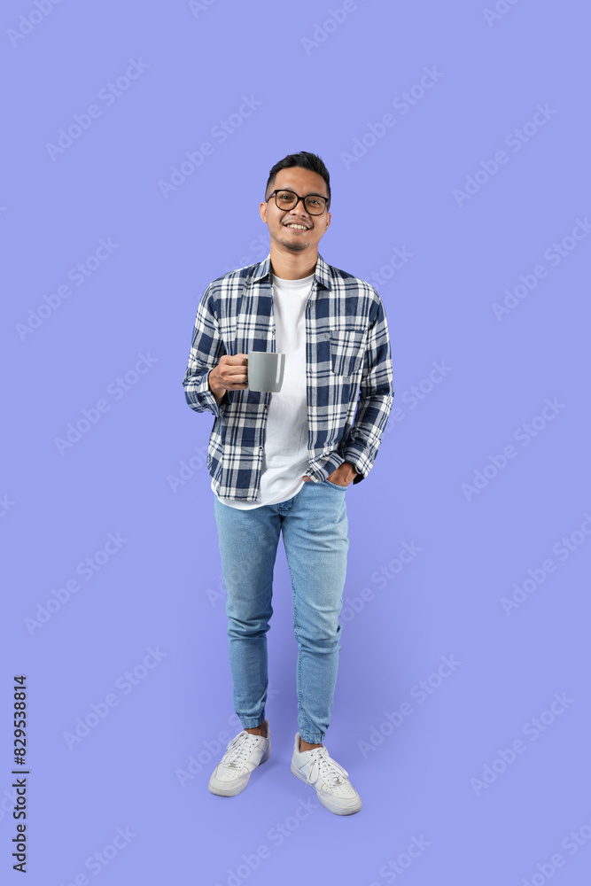 Fototapeta premium Full-length Portrait of a young Asian man smiling while holding a cup standing isolated over a purple background