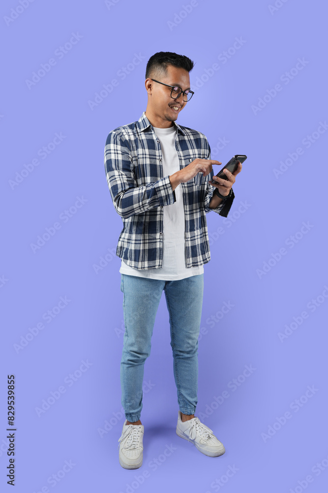 Full-length Portrait of a young cheerful excited Asian man standing isolated over a purple background, using a mobile phone Full-length Portrait of a young cheerful excited Asian man standing isolated over a purple background, using a mobile phone