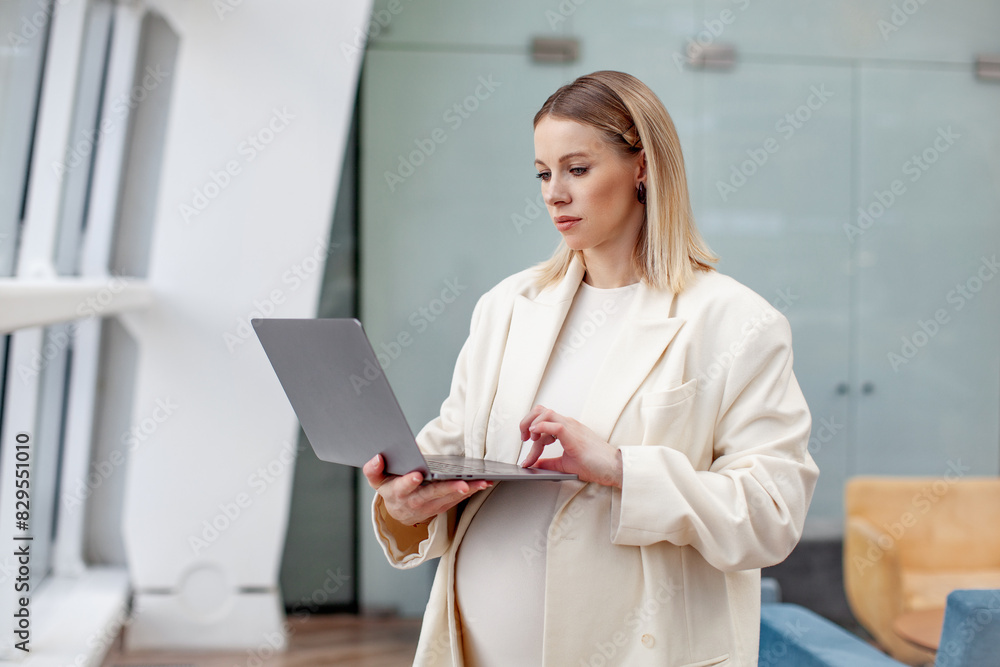 Portrait of beautiful pregnant businesswoman confidently working on laptop in a modern business office with a light blue background, wearing a stylish suit and blazer, representing female empowerment Portrait of beautiful pregnant businesswoman confidently working on laptop in a modern business office with a light blue background, wearing a stylish suit and blazer, representing female empowerment
