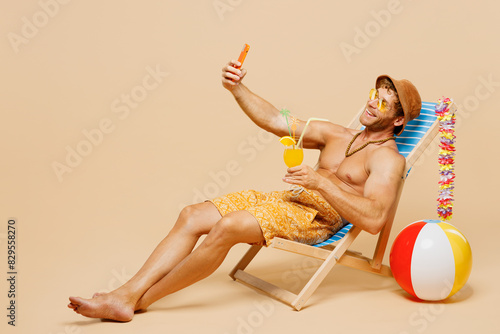Full body young man wear yellow shorts swimsuit relax rest near hotel pool sit in deckchair do selfie shot on mobile cell phone isolated on plain beige background. Summer vacation sea sun tan concept. © ViDi Studio