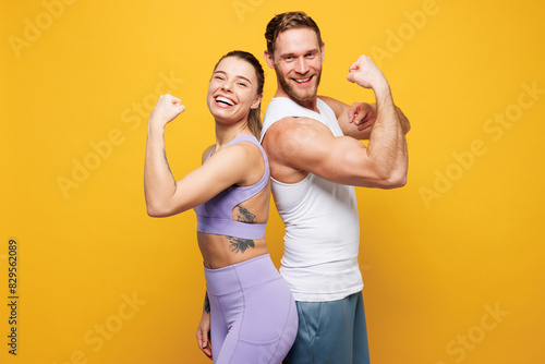 Young strong fitness trainer two man woman wearing blue clothes spend time in home gym showing biceps muscles on hand stand back to back isolated on plain yellow background. Workout sport fit concept.