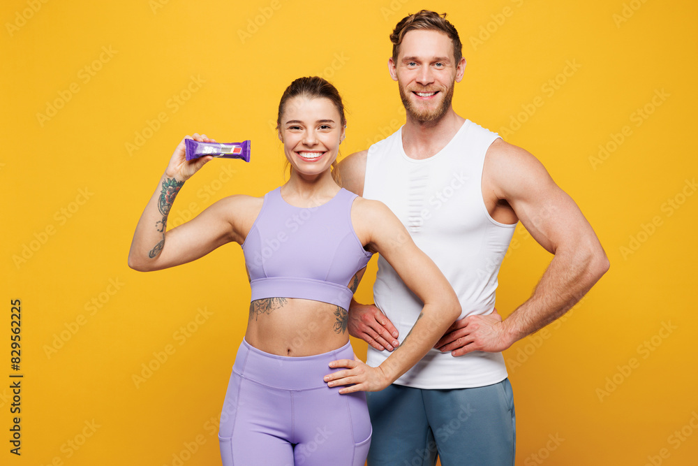 Young strong fitness trainer instructor sporty two man woman wears blue clothes spend time in home gym hold in hand protein energy bar isolated on plain yellow background. Workout sport fit concept. Young strong fitness trainer instructor sporty two man woman wears blue clothes spend time in home gym hold in hand protein energy bar isolated on plain yellow background. Workout sport fit concept.
