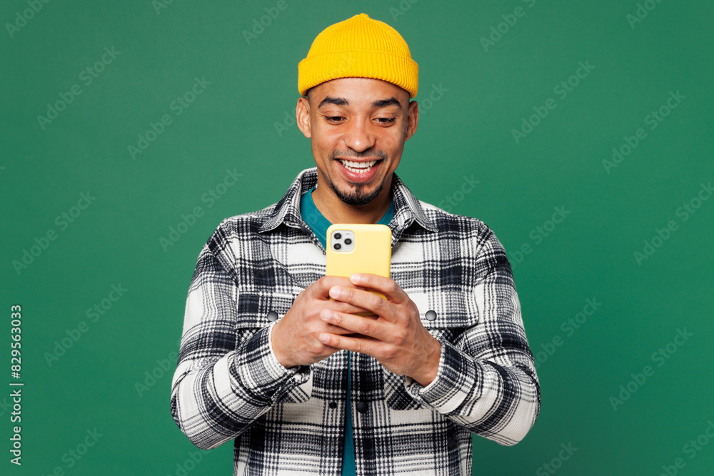 Young man of African American ethnicity he wear shirt blue t-shirt yellow hat hold in hand use mobile cell phone chatting online isolated on plain green background studio portrait. Lifestyle concept. Young man of African American ethnicity he wear shirt blue t-shirt yellow hat hold in hand use mobile cell phone chatting online isolated on plain green background studio portrait. Lifestyle concept.