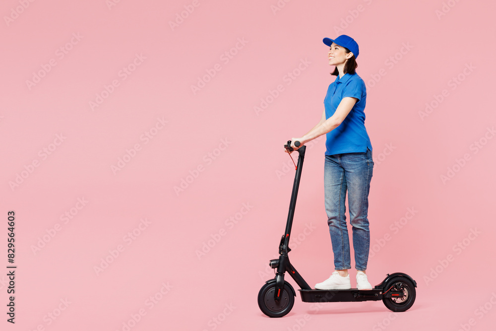 Full body side view professional delivery girl employee woman wears blue cap t-shirt uniform workwear work as dealer courier ride e-scooter isolated on plain pastel pink background. Service concept.