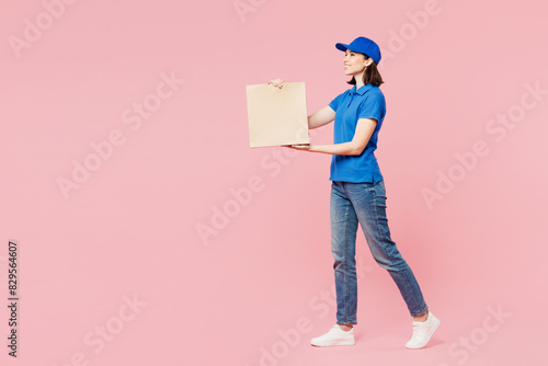 Full body side view delivery girl employee woman wear blue cap t-shirt uniform workwear work as dealer courier hold brown blank craft paper bag go isolated on plain pink background. Service concept. photo