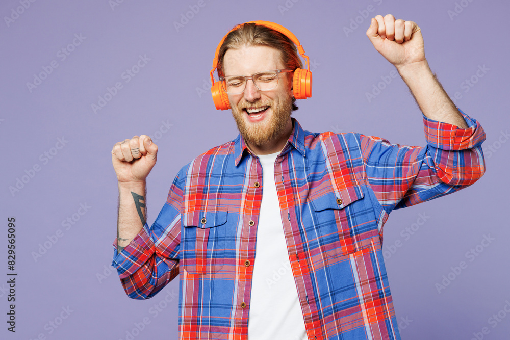 Naklejka premium Young smiling fun happy man he wears blue shirt casual clothes listen to music in headphones raise up hands dance isolated on plain pastel light purple background studio portrait. Lifestyle concept.
