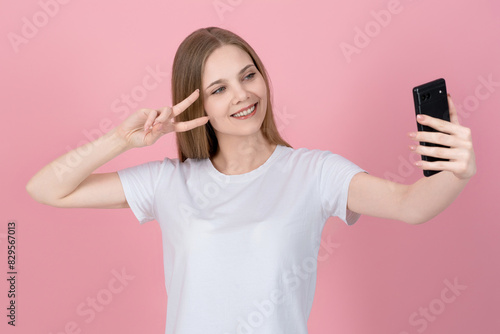 Attractive caucasian young blonde woman in casual white t-shirt taking selfie on mobile phone camera, showing peace gesture isolated on pink studio background.