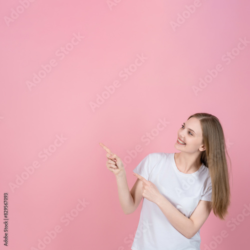 Attractive caucasian young blonde woman in casual white t-shirt smiling and pointing fingers at empty copy space for advertising on pink studio background. Sale, advice, choice.