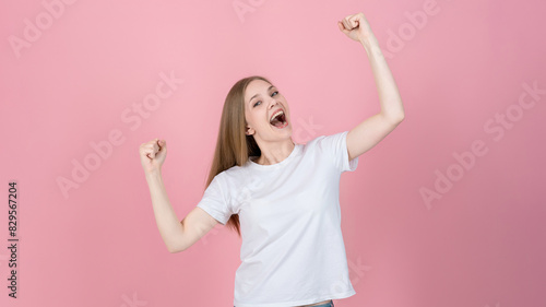 Attractive caucasian young blonde woman in casual white t-shirt looking at camera, smiling happily and showing winner gesture isolated on pink studio background. Success, victory.