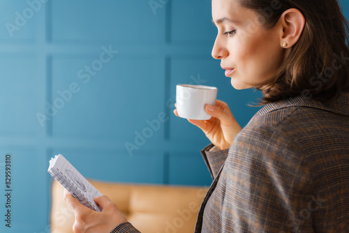 Beautiful businesswoman is standing in the office, reading notes from a notepad and drinking coffee.