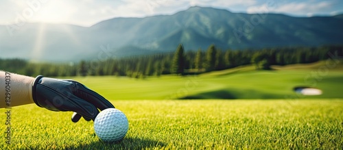 A hand in a glove sets a golf tee with a ball in the ground on a golf course featuring lush green grass and mountains in the backdrop with a soft focus creating a blurred effect ideal for a copy spac photo