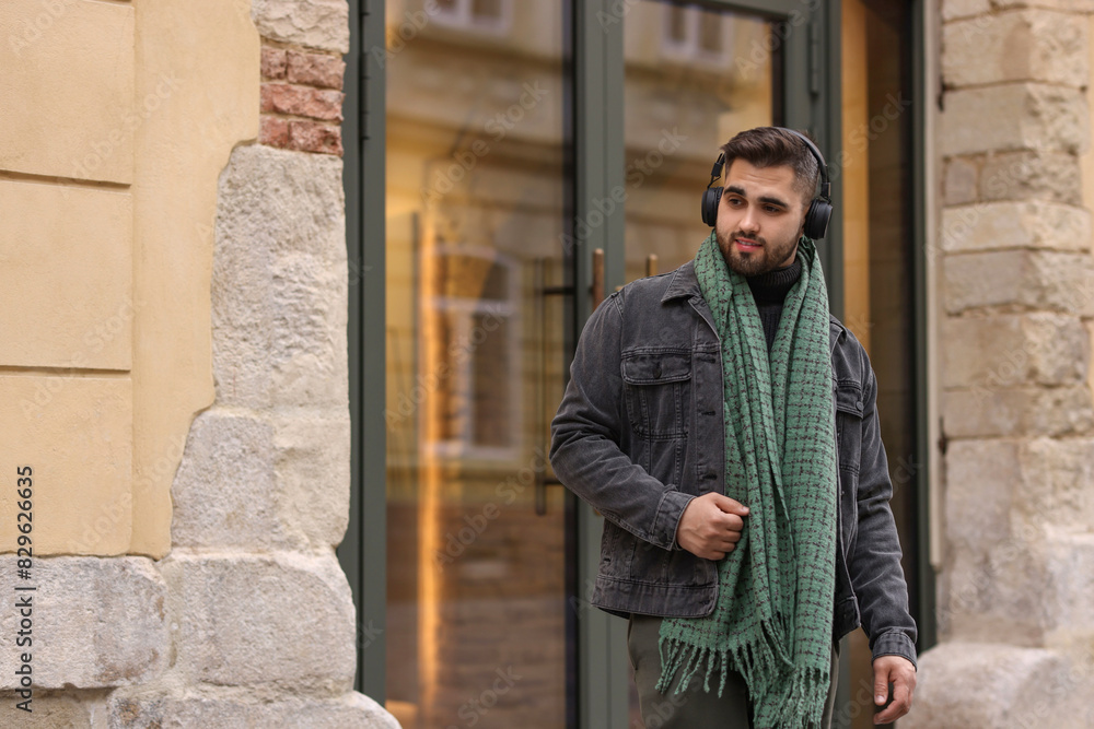 Handsome man in warm scarf and headphones near building outdoors. Space for text Handsome man in warm scarf and headphones near building outdoors. Space for text
