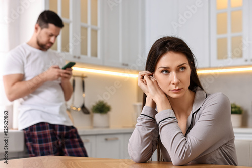 Offended wife sitting at table while her husband using smartphone in kitchen, selective focus. Relationship problems photo