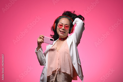 Cheerful elderly woman with headphones enjoying music dancing over pink background © Prathankarnpap