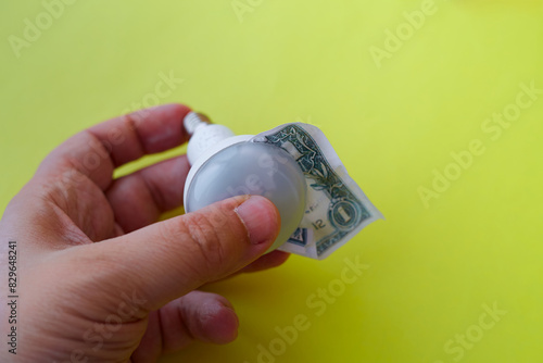 A hand holds an LED bulb from which a dollar peeks out on a yellow background, saving on electricity, economical technologies, payment of utility bills