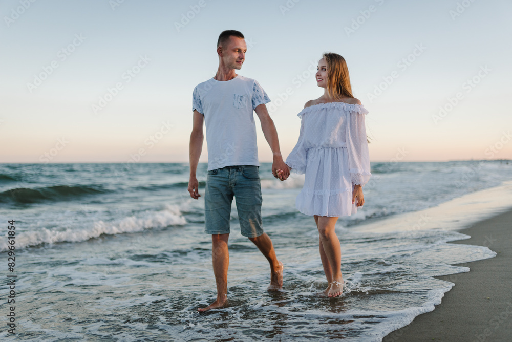 Man and woman walking on water with big waves in sea. Happy couple in love holding hands, looking at each other walking barefoot on seashore. Female and male run on sand beach ocean sunny summer day.