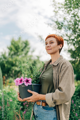 Beautiful adult redhead woman in her garden holding flower pot with petunia wearing casual style and short haircut wellness and nature calm