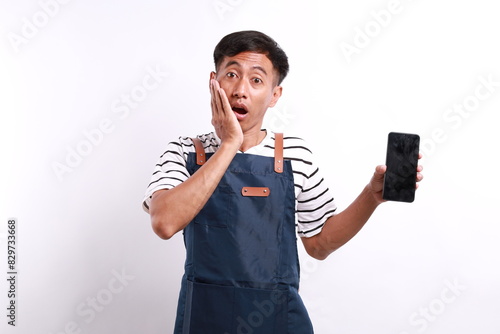 Shocked amazed young asian man barista bartender barman employee in apron hold mobile phone with blank empty screen mock up copy space put hand on cheek isolated on white background studio portrait photo