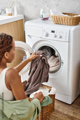 An African American woman with afro braids carefully loads clothes into a washing machine in a bathroom. photo