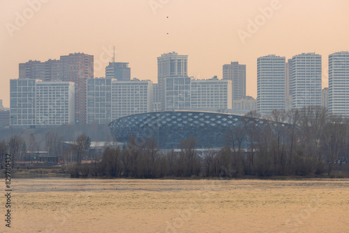Evening cityscape. View of the sports ice palace and modern residential high-rise buildings. Novosibirsk city, Novosibirsk region, Siberia, Russia. In the foreground is the Ob River. Big Siberian city