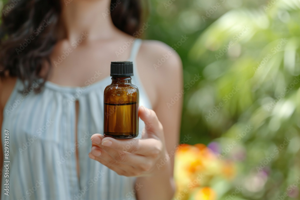 Fototapeta premium A woman is holding a bottle of essential oil. The bottle is brown and has a black cap. The woman is standing in a garden with trees and flowers in the background. Concept of relaxation and tranquility