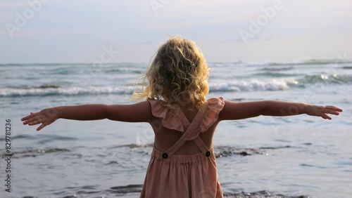 Little girl in dress stands with her arms wide open on beach, enjoying wind and looking at waves sea. Powerful waves crash with crash on stones sea near child. Sea in pristine beauty and power