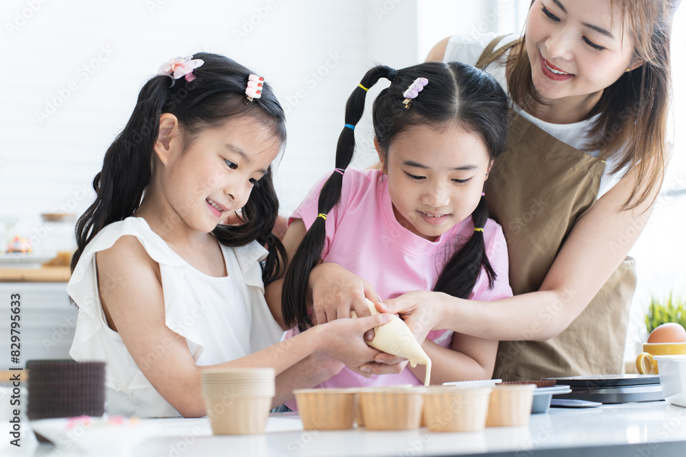 Asian family, mother and two daughters enjoy making cupcakes together at home kitchen. Mom and little child girl squeezing cake batter into cupcake paper cup, prepare for bake, smiling. Happy holiday Asian family, mother and two daughters enjoy making cupcakes together at home kitchen. Mom and little child girl squeezing cake batter into cupcake paper cup, prepare for bake, smiling. Happy holiday