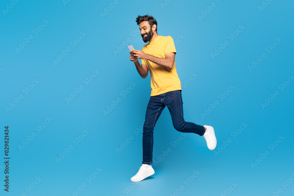 Fototapeta premium An adult Indian man wearing a yellow shirt is jogging outdoors with his attention focused on a smartphone screen.