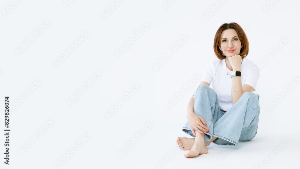 Attractive middle-aged woman in jeans and barefoot on a white background. Portrait of a woman in casual clothes. A banner with an empty space for text