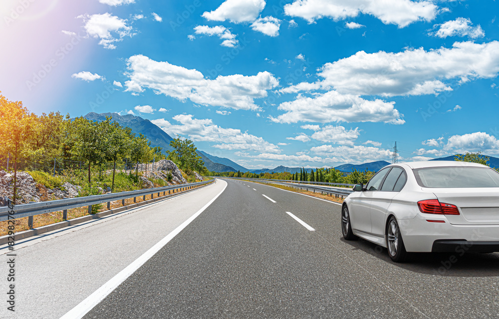 A white car drives along the highway against the backdrop of rocky mountains on a sunny day.