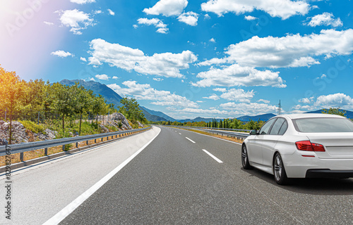 A white car drives along the highway against the backdrop of rocky mountains on a sunny day.