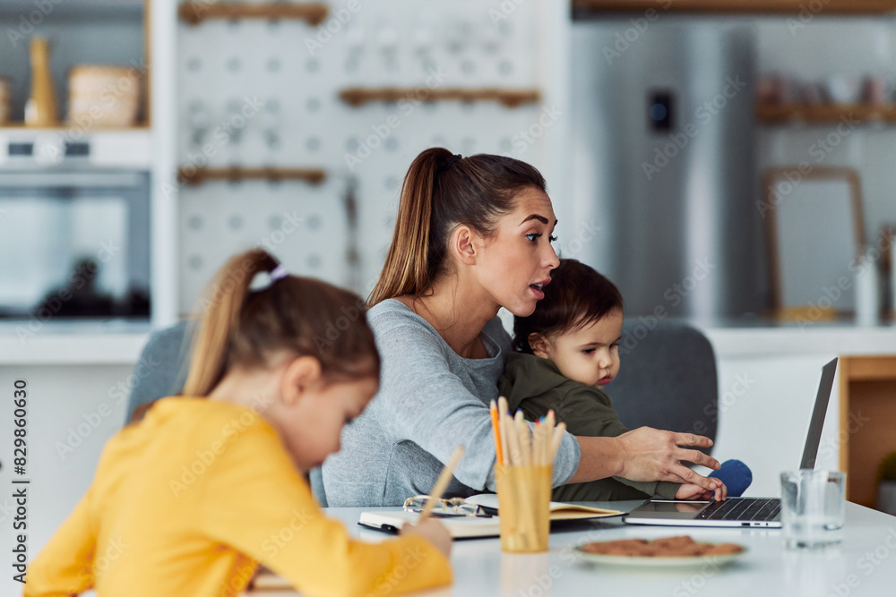 Fototapeta premium A hard-working mother having a video call on her laptop while working from home and looking after her kids