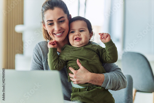 A happy mother holding her baby while working on her laptop from home photo