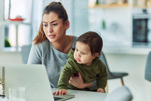 A chagrined mother holding her baby as the child curiously touches the laptop and disrupts mother's work making her feel stressed photo