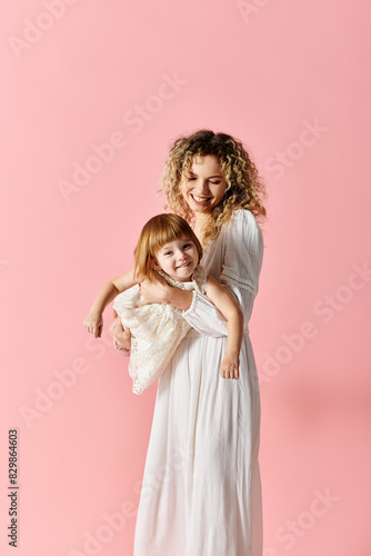 A mother with curly hair holds her baby on a pink background.